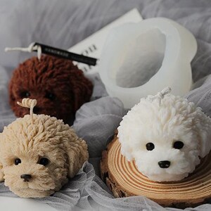 May include: A collection of dog-shaped candles in various colors, including brown, beige, and white. One candle is placed on a wooden slice. A silicone mold is also visible. The candles have detailed features and are set against a gray fabric background.