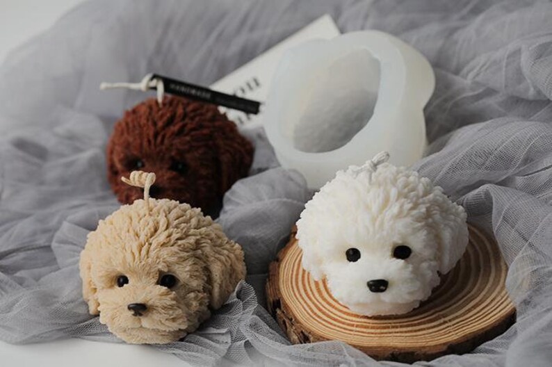 May include: A collection of dog-shaped candles in various colors, including brown, beige, and white. One candle is placed on a wooden slice. A silicone mold is also visible. The candles have detailed features and are set against a gray fabric background.
