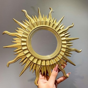 May include: A gold-colored sunburst mirror with a round central mirror. The sunburst design features pointed rays and curved accents. The mirror is held up by a hand with painted nails.