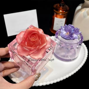 Puede incluir: Una caja de cristal rosa y cuadrada con una gran rosa roja encima. La caja está sobre un plato ovalado blanco, junto a un pequeño tarro de cristal morado con una flor. El texto "Daisy Note Craft" es visible.