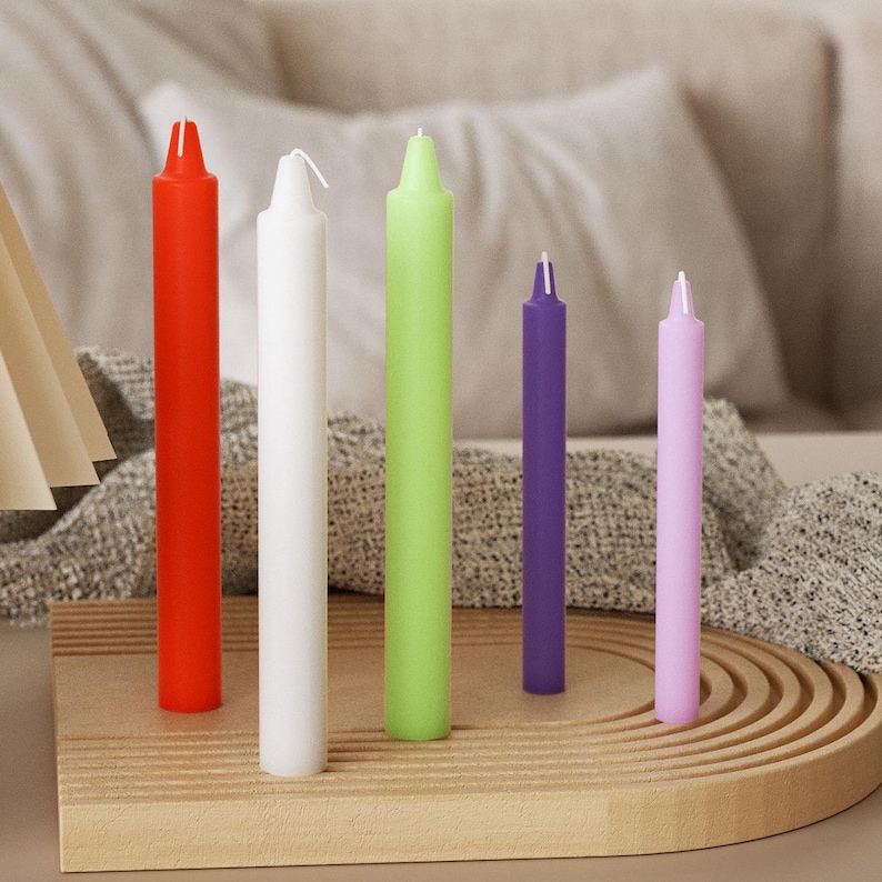 May include: Five colorful taper candles in red, white, green, purple, and pink are arranged on a wooden tray. The candles vary in height and are unlit, with visible wicks. The background includes a neutral-toned blanket and pillows.