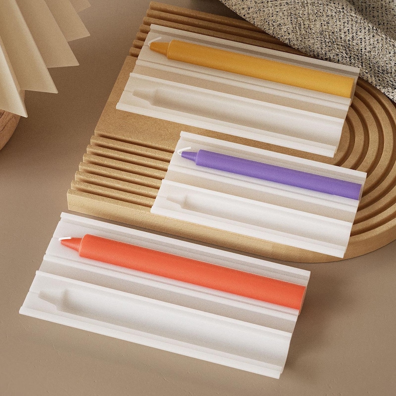 May include: Three colorful candles in white rectangular holders. The candles are orange, purple, and yellow. The holders are on a wooden tray with a ridged design. The background includes a beige fabric and a light-colored surface.