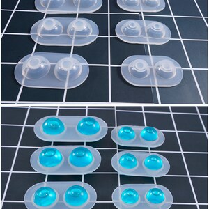 May include: Clear silicone molds with two rounded cavities each. Some molds contain blue, dome-shaped objects. The molds are arranged on a black and white grid surface. The molds are likely for crafting or resin art.