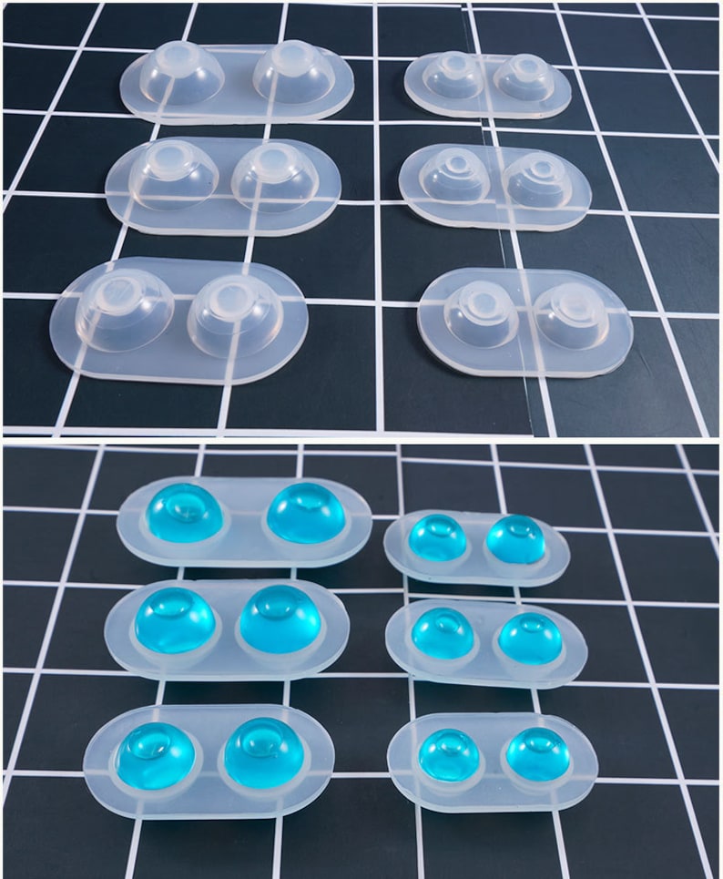 May include: Clear silicone molds with two rounded cavities each. Some molds contain blue, dome-shaped objects. The molds are arranged on a black and white grid surface. The molds are likely for crafting or resin art.