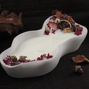 May include: A white candle in a decorative ceramic dish with a wavy edge. The candle is decorated with dried flowers, cinnamon sticks, and dried orange slices. The candle is surrounded by acorns and a dried leaf.
