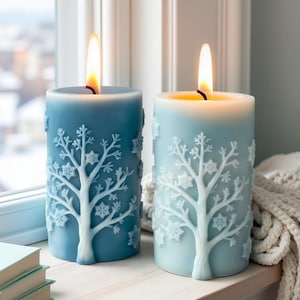 Puede incluir: Dos velas de pilar azules encendidas con un diseño de árbol de invierno y copos de nieve. Las velas son de diferentes tonos de azul y están sobre una superficie de madera cerca de una ventana. Las llamas son de color amarillo brillante.