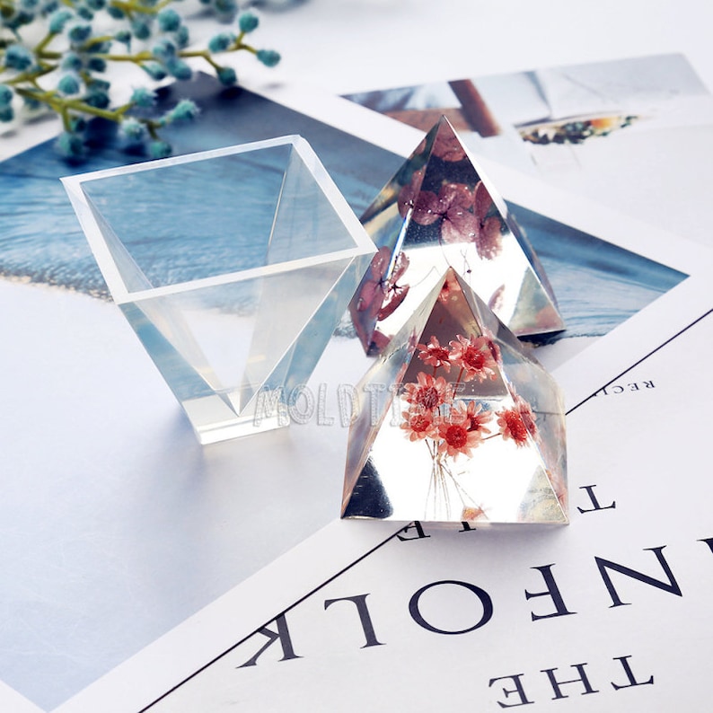 May include: Clear resin pyramid and square molds. Two pyramids contain dried red flowers. The square mold is empty. The objects are on a white surface with text and a sprig of blue flowers.