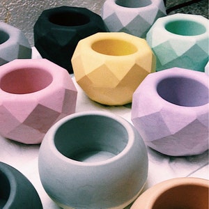 Puede incluir: Una colección de macetas geométricas facetadas en varios colores pastel, incluyendo rosa, amarillo, verde y morado. Las macetas tienen forma redonda con una amplia abertura, adecuadas para pequeñas plantas o elementos decorativos.