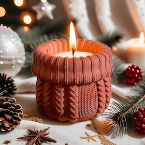 Könnte beinhalten: Eine brennende Kerze in einem terrakottafarbenen Kerzenhalter im Strickstil. Die Kerze hat einen weißen Wachsinnenraum und eine flackernde Flamme. Der Halter hat ein Zopfmuster. Der Hintergrund umfasst Weihnachtsdekorationen und Tannennadeln.