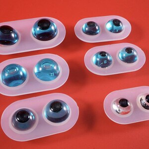 May include: Several pairs of glass eyes in various shades of blue and silver, each with a black pupil. The eyes are set in clear, oval-shaped molds, arranged against a solid red background. The eyes vary in size and color.