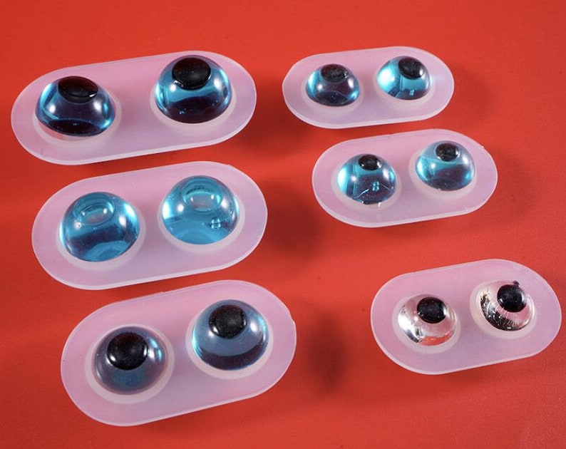 May include: Several pairs of glass eyes in various shades of blue and silver, each with a black pupil. The eyes are set in clear, oval-shaped molds, arranged against a solid red background. The eyes vary in size and color.