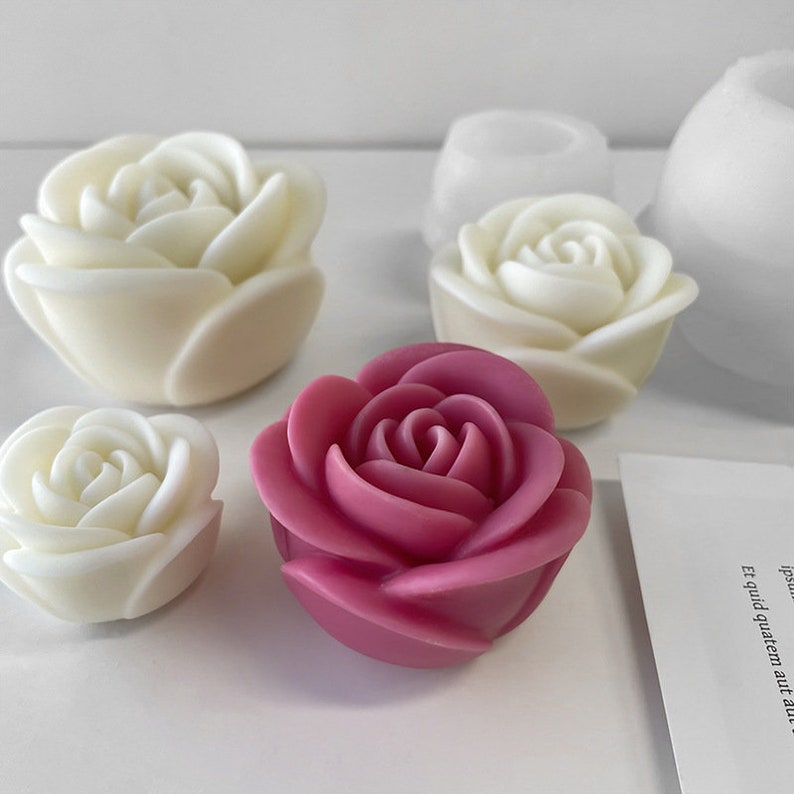May include: A selection of rose-shaped candles in white and deep pink. The candles are arranged on a white surface. The image also includes a white paper with text. The candles are likely made of wax.