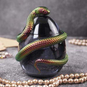 May include: A dark glass jar with a lid, adorned with a metallic green and red iridescent snake. The snake's head is on the lid. The jar is egg-shaped and rests on a grey surface.