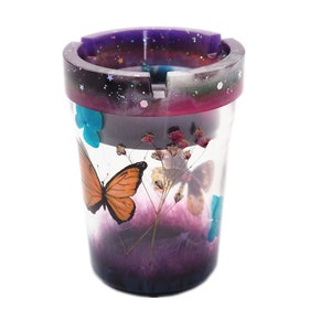 Puede incluir: Cenicero cil&iacute;ndrico con cuerpo transparente, con una mariposa naranja y flores secas. El borde tiene un degradado p&uacute;rpura y negro con purpurina. Tambi&eacute;n se ven acentos de mariposas azules.