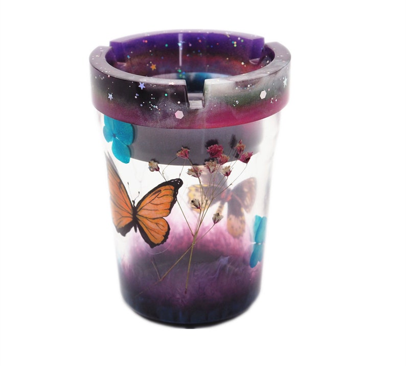 Puede incluir: Cenicero cil&iacute;ndrico con cuerpo transparente, con una mariposa naranja y flores secas. El borde tiene un degradado p&uacute;rpura y negro con purpurina. Tambi&eacute;n se ven acentos de mariposas azules.