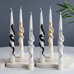 Puede incluir: Velas blancas cónicas adornadas con adornos decorativos de serpientes en negro, dorado y plateado. Las serpientes se enrollan alrededor de las velas, agregando un toque único y artístico. Las velas están dispuestas sobre una superficie de tono neutro.