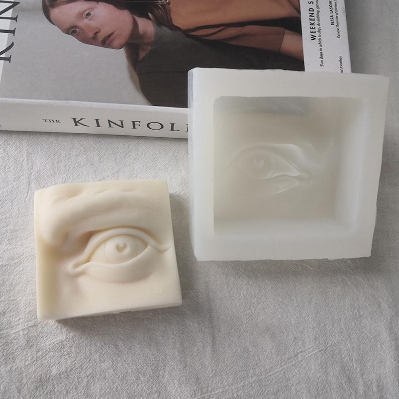 May include: A square, off-white soap bar with a sculpted eye design next to a white silicone mold with the same eye shape. The soap has a detailed, three-dimensional eye, while the mold has a recessed eye shape. A book is in the background.