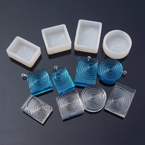 Pode incluir: Uma coleção de moldes de resina transparentes e azuis e pingentes acabados. Os moldes são retangulares e circulares, com um design de círculos concêntricos. Os pingentes têm uma argola prateada.