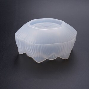 Puede incluir: Un molde de silicona transparente para manualidades. El molde tiene una forma redondeada, similar a un cuenco, con un exterior texturizado y ondulado y una parte superior plana. El borde inferior tiene un dise&ntilde;o festoneado.