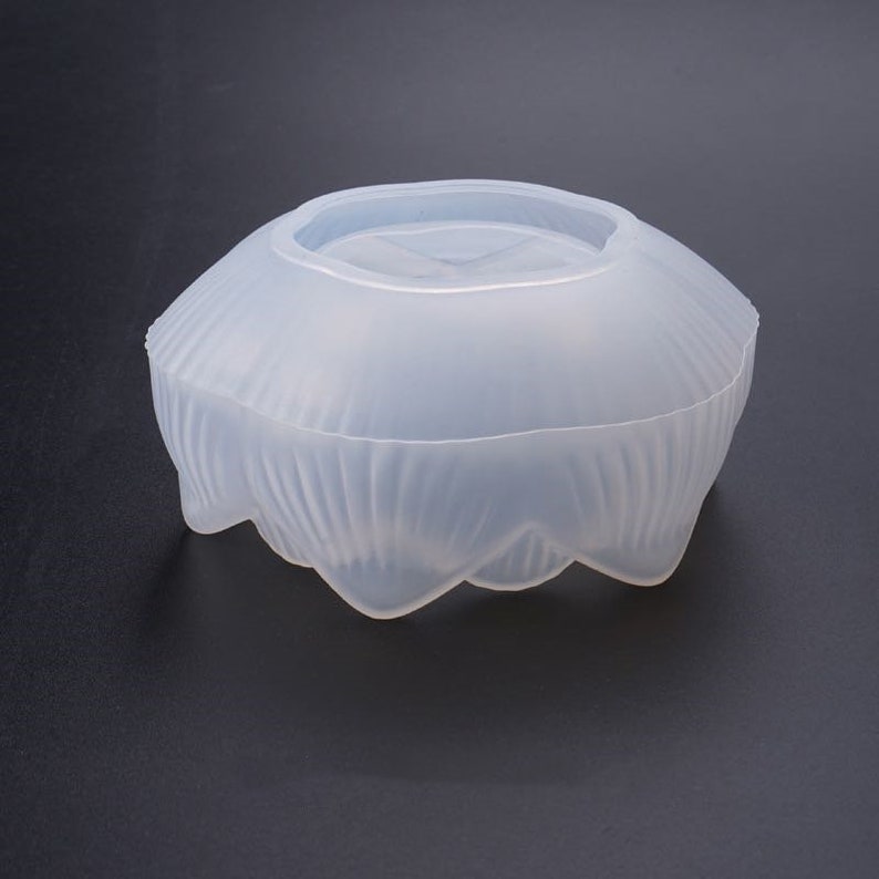 Puede incluir: Un molde de silicona transparente para manualidades. El molde tiene una forma redondeada, similar a un cuenco, con un exterior texturizado y ondulado y una parte superior plana. El borde inferior tiene un dise&ntilde;o festoneado.