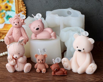 Puede incluir: Una colección de velas con forma de osito de peluche en varios tamaños y tonos de rosa y marrón. Algunos osos están sentados, tumbados o sosteniendo osos más pequeños. Hay moldes de velas blancas en el fondo. Cada vela tiene una pequeña etiqueta.