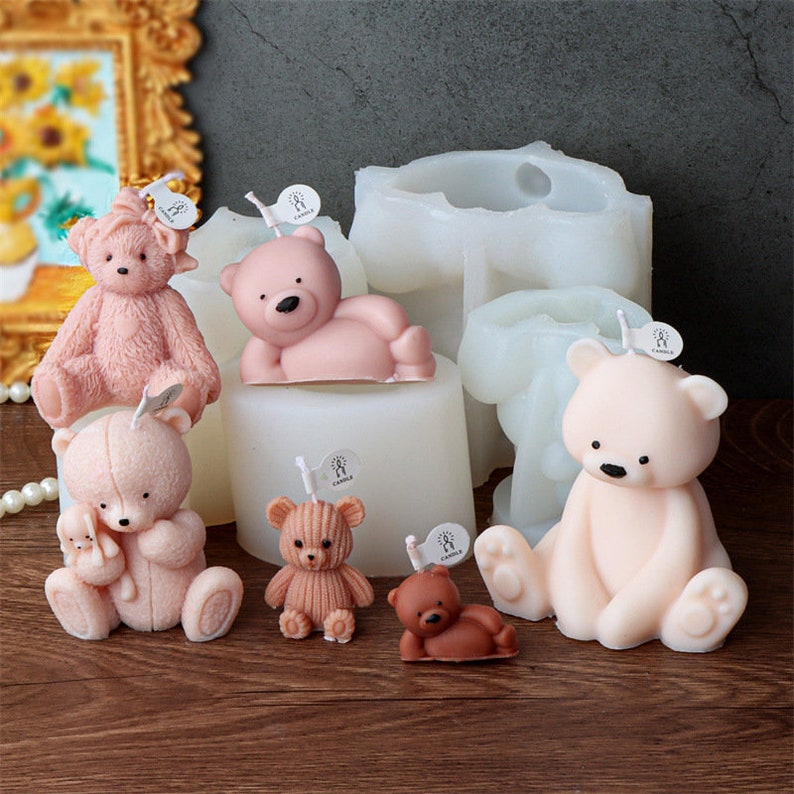 May include: A collection of teddy bear-shaped candles in various sizes and shades of pink and brown. Some bears are sitting, lying down, or holding smaller bears. White candle molds are in the background. Each candle has a small tag.