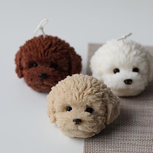 May include: Three dog-shaped candles in varying colors: brown, cream, and white. Each candle has a textured, fluffy appearance, with black eyes and noses. The candles are arranged on a neutral surface.