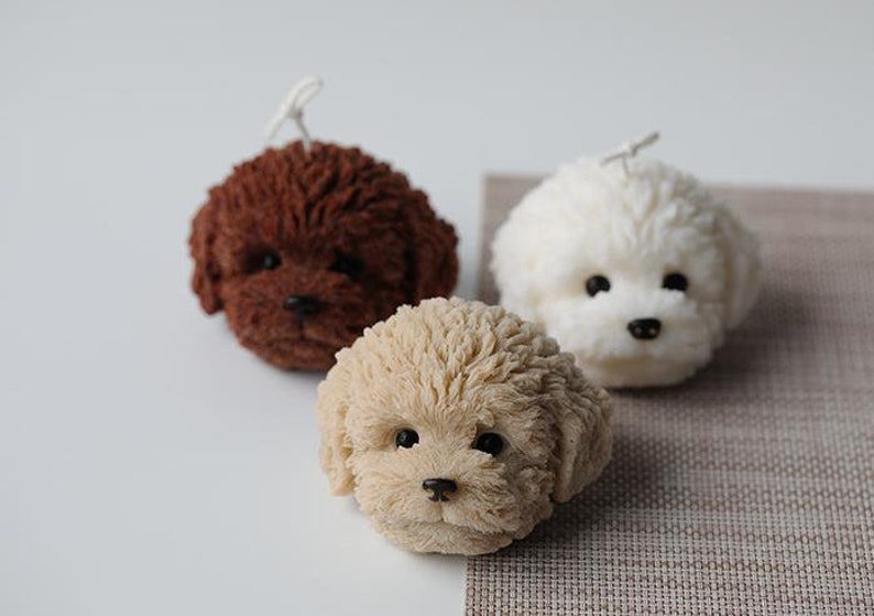 May include: Three dog-shaped candles in varying colors: brown, cream, and white. Each candle has a textured, fluffy appearance, with black eyes and noses. The candles are arranged on a neutral surface.