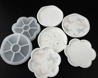 May include: An assortment of white resin trays and molds in various shapes, including round and flower-shaped designs. Some trays feature butterfly patterns and speckled details. The items are displayed against a black background.