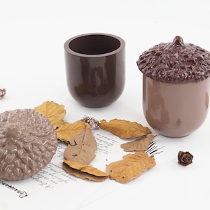 May include: A collection of ceramic acorn-shaped containers and a cylindrical container in shades of brown. The acorn-shaped pieces have textured caps. Dried leaves and small pine cones are scattered around the containers on a white surface.
