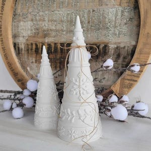 Puede incluir: Dos adornos blancos de cerámica con forma de árbol de Navidad con diseños de copos de nieve y hojas. El árbol más grande está atado con hilo. Los árboles están sobre un fondo de madera rústica con ramas de algodón.