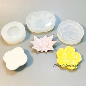 Könnte beinhalten: Eine Auswahl an Bastelartikeln, darunter transparente Silikonformen in verschiedenen Formen: ein Kreis, ein Ahornblatt und eine Blume. Es gibt auch fertige Resin-Teile in Rosa und Gelb sowie den Text "DaisyNoteCraft".