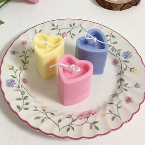 May include: Three heart-shaped candles in pastel colors: yellow, pink, and blue. Each candle has a white wick and sits on a decorative plate with floral patterns. The candles are a charming decorative item.