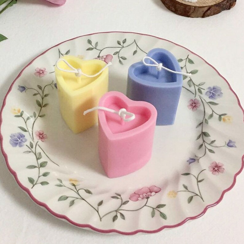May include: Three heart-shaped candles in pastel colors: yellow, pink, and blue. Each candle has a white wick and sits on a decorative plate with floral patterns. The candles are a charming decorative item.