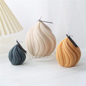 May include: Three decorative candles in a swirl design. The candles are in shades of beige, blue-grey, and yellow-orange. Each candle has a wick and a small black tag that reads "HANDMADE". The candles are on a white surface.