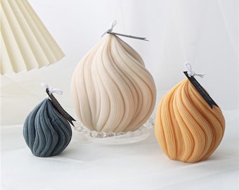 May include: Three decorative candles in a swirl design. The candles are in shades of beige, blue-grey, and yellow-orange. Each candle has a wick and a small black tag that reads "HANDMADE". The candles are on a white surface.