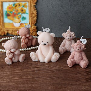 May include: Five teddy bear-shaped candles in varying shades of pink and brown are arranged on a wooden surface. Each candle has a wick and a small tag. The bears have black eyes and are in different poses.