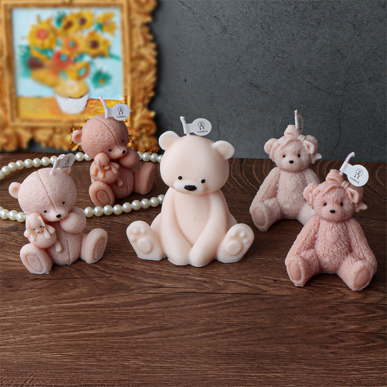 May include: Five teddy bear-shaped candles in varying shades of pink and brown are arranged on a wooden surface. Each candle has a wick and a small tag. The bears have black eyes and are in different poses.