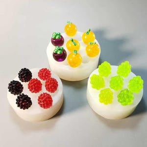 Op de afbeelding: Drie witte, ronde objecten, elk bekroond met kleurrijk miniatuurfruit. Eén heeft rode en zwarte bessen, een ander gele sinaasappels en paarse mangosteen, en de laatste groene druiven. Het fruit is gerangschikt op een witte basis.