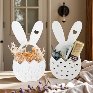 May include: Two white Easter-themed decorative baskets shaped like bunny heads. One basket contains dried orange flowers, and the other holds a small gift. The word "Ostern" is carved on the front of one basket.