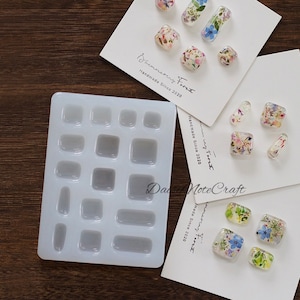 May include: A clear silicone mold with various square and rectangular cavities, alongside several resin pieces embedded with dried flowers. The resin pieces are in various shapes, including squares and oblongs. The text "Shimmering Foret Handmade Since 2020" is visible.