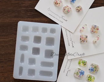 May include: A clear silicone mold with various square and rectangular cavities, alongside several resin pieces embedded with dried flowers. The resin pieces are in various shapes, including squares and oblongs. The text "Shimmering Foret Handmade Since 2020" is visible.