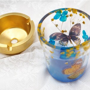 Puede incluir: Un cenicero dorado con tres ranuras y un agujero central se encuentra junto a una copa de cristal. La copa est&aacute; decorada con detalles azules y amarillos y presenta un dise&ntilde;o de mariposa y floral. La copa est&aacute; llena de un l&iacute;quido azul.