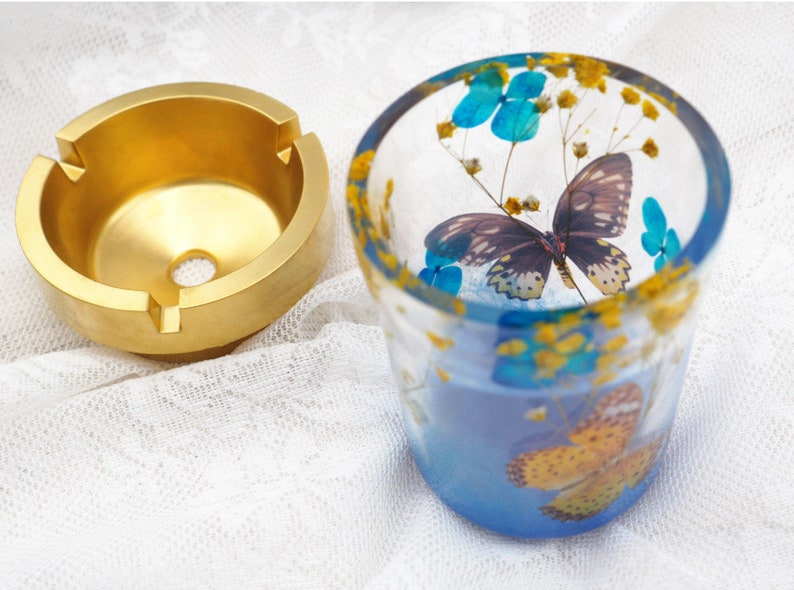 Puede incluir: Un cenicero dorado con tres ranuras y un agujero central se encuentra junto a una copa de cristal. La copa est&aacute; decorada con detalles azules y amarillos y presenta un dise&ntilde;o de mariposa y floral. La copa est&aacute; llena de un l&iacute;quido azul.