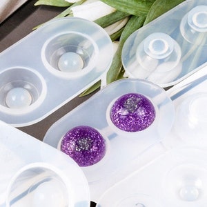 May include: Close-up of several clear silicone molds, each with circular cavities. Some molds contain spherical objects, including a purple glitter sphere. The molds are designed for crafting and creating resin art.