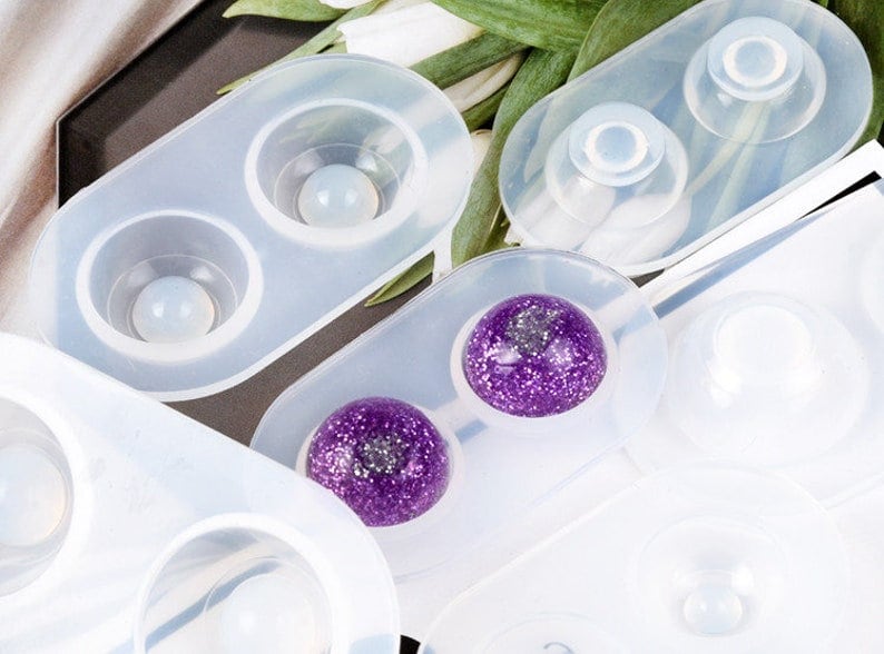 May include: Close-up of several clear silicone molds, each with circular cavities. Some molds contain spherical objects, including a purple glitter sphere. The molds are designed for crafting and creating resin art.