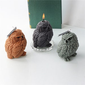 May include: Three owl-shaped candles in shades of brown, gray, and dark gray. The dark gray candle has a lit wick. Each candle has a textured surface and is labeled with a small tag. The candles are on a white surface.