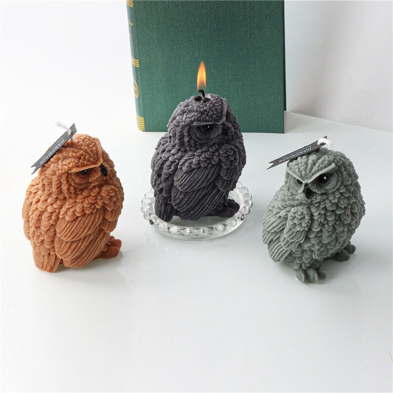 May include: Three owl-shaped candles in shades of brown, gray, and dark gray. The dark gray candle has a lit wick. Each candle has a textured surface and is labeled with a small tag. The candles are on a white surface.