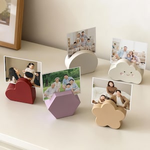 Op de afbeelding: Een verzameling fotohouders in verschillende vormen: een hart, een zeshoek, een wolk, een bloem en een halve cirkel. Elke houder toont een familiefoto. De houders zijn in tinten rood, lavendel, beige en wit.