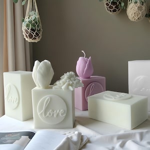 Puede incluir: Un surtido de velas decorativas de varias formas y colores. Las velas incluyen velas blancas y lavanda con forma de tulipán, y velas cúbicas blancas con la palabra "love" en relieve. Las velas están dispuestas sobre una superficie blanca.